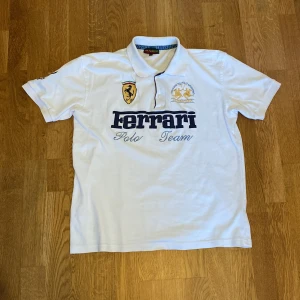 Vit Ferrari Polo Team piké med märket La Martina  - Vit t-shirt från La Martina i klassisk polomodell med broderade Ferrari Polo Team-loggor fram och bak. Snygg krage, korta ärmar och detaljer i blått och guld. Tillverkad i mjuk bomull. Det står storlek XL men den har krympt vilket gör att den nu passar M/L bättre. Den enda defekten på tröjan är 4 små hål på sidan av tröjan där de knappt syns, den största har 1 mm i diameter.
