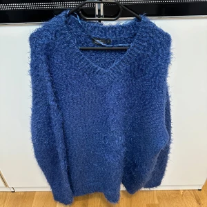 Blå fluffig stickad tröja  - Mysig blå stickad tröja från KappAhl med fluffig textur och v-ringad hals. Perfekt för kyliga dagar. Långärmad modell som ger en avslappnad och trendig look.