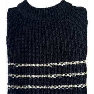 Svart stickad tröja med vita ränder och en liten öppning i slutet  - Svart stickad tröja med silvriga horisontella ränder och grov ribbstickad struktur. Tröjan har rund halsringning och långa ärmar. Perfekt för dig som gillar en klassisk men ändå lite extra look.