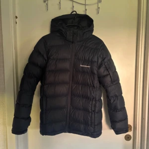 Svart dunjacka Peak Performance 170 - Svart dunjacka från Peak Performance med huva och dragkedja framtill. Jackan har quiltad design och är tillverkad i slitstark polyester med Pertex Quantum-tyg. Perfekt för kalla dagar och har en clean, sportig look. ‼️BARA ANVÄND EN VINTER) 