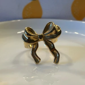 Guldig ring med rosett - Superfin ring i guldton med en stor, glänsande rosett framtill. Ringen har öppen baksida så den är justerbar och passar flera storlekar. Perfekt accessoar för att ge din look en söt och trendig touch.