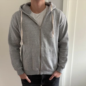 Grå hoodie med dragkedja och huva - Grå hoodie med dragkedja framtill, huva med vita snören och två fickor. 