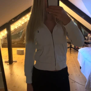 Vit hoodie från Polo Ralph Lauren - Vit hoodie från Polo Ralph Lauren med dragkedja framtill och klassisk blå logga på bröstet. Tröjan har huva, känguruficka och är tillverkad i mjuk bomull. Perfekt för en chill och stilren look.