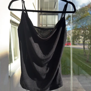 Svart linnetopp med draperad urringning från Gina  - Säljer en svart linnetopp med smala axelband och snyggt draperad urringning. Toppen är figurnära och har en stilren, enkel design som passar perfekt till jeans eller kjol. Materialet känns mjukt och följsamt mot kroppen. Och kommer från Gina 