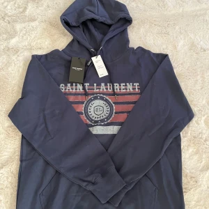 Blå Saint Laurent hoodie med tryck - Säljer en riktigt snygg och stilren Saint Laurent hoodie i marinblått, storlek M. Hoodien är i nyskick (10/10) och endast testad. Modellen har en retroinspirerad logga framtill med klassiska ”Saint Laurent”-trycket.  Nypris ca 8000 kr – vårt pris: 1999 kr  Tveka inte att höra av dig vid frågor! 🙌.