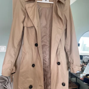 Beige trenchcoat från Cubus, storlek XS - Stilren beige trenchcoat från C i storlek XS. Jackan har klassisk dubbelknäppning, bälte i midjan och snygga detaljer vid ärmsluten. Tillverkad i ett lätt bomullsmaterial, perfekt för övergångssäsonger. Snygg krage och fickor på sidorna.