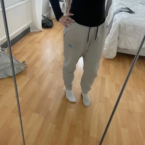 Beige mjukisbyxor från Nike M - Säljer ett par beige nike tech byxor från Nike i storlek M. Byxorna har svarta dragsnören i midjan, svarta dragkedjor på fickorna och en blå Nike-logga på ena benet. Sköna med elastisk midja och muddar vid bensluten. Det finns en fläck av bläck på framsidan av byxan som inte går bort men är inte jätte synlig.
