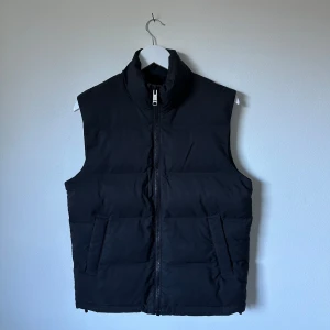 Svart dunväst från Weekday XS - Svart dunväst från Weekday i storlek XS. Västen har hög krage, två fickor framtill och dragkedja hela vägen. Perfekt att slänga över en hoodie för extra värme. Enkel, stilren och passar till många olika looks. 3Y19