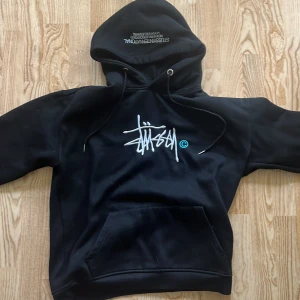 Svart Stussy hoodie med brodyr, M - Svart hoodie från Stussy med vit broderad logga på bröstet och text på huvan. Klassisk känguruficka, snörning i huvan och mjukt material. Perfekt för dig som gillar streetwear och vill ha en stilren look.
