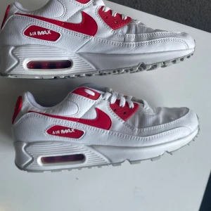 Nike Air Max 90 vit/röd sneakers - Nike Air Max 90 sneakers i vitt och rött med klassisk synlig Air Max-dämpning i sulan. Skorna har röda detaljer och swoosh, vit mesh och skinnpaneler samt snörning. Perfekt för dig som gillar retrostil och vill sticka ut med en ikonisk modell.