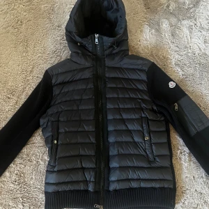 Svart Moncler cardigan - Säljer en svart cardigan från Moncler med stickade ärmar och huva. Storlek L men passar M då dom är små i storleken. Jackan har dragkedja framtill, två fickor med knappar och en extra ficka med dragkedja på ärmen. Klassisk Moncler-logga på vänster ärm. Perfekt för kyliga dagar. NFC scan, frågor? Kontakta mig 