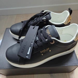 Svarta sneakers från Replay strl 38 - Snygga svarta sneakers från Replay med gulddetaljer och vit sula. Skorna har snörning, Replay-logga i guld på sidan och hälparti i metallicguld. Ovandel i skinnliknande material och insida i vitt. Perfekta för en trendig vardagslook.