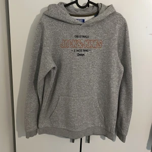 Grå hoodie från Jack & Jones - Grå hoodie från Jack & Jones med broderad logga och text på bröstet. Tröjan har huva, magficka och långa ärmar. Mjuk insida och klassisk passform, perfekt för chill dagar. Snygg och enkel att matcha med jeans eller joggers.