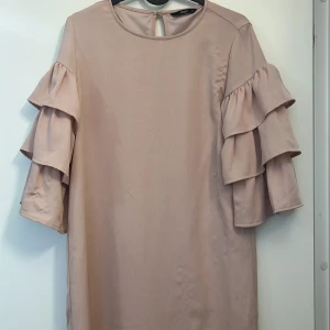 Beige volangklänning från ONLY - Supersöt ljusrosa kortklänning från ONLY med rund halsringning och trekvartsärmar med dubbla volanger. Klänningen har en rak passform och är tillverkad i ett mjukt, lätt material som känns skönt mot huden. Perfekt för dig som gillar en stilren look med en twist.