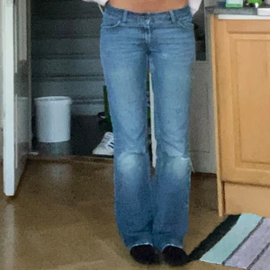 Blå bootcut jeans med hål vid knäna och slitningar - Säljer ett par vintage blå bootcut jeans med snygga slitningar vid knäna och lätt tvättade detaljer. Jeansen har låg midja och klassisk femficksdesign. 