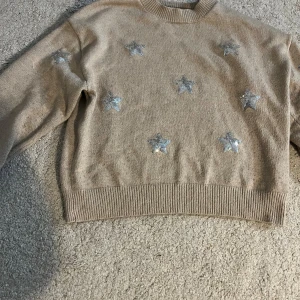 Beige stickad tröja med silverstjärnor - Beige stickad tröja från Gina Tricot med glittriga silverstjärnor framtill. Tröjan har rund hals, ribbade muddar och lång ärm. Perfekt för dig som gillar lite extra bling i vardagen.