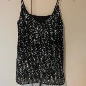 Svart paljettlinne med smala axelband - Glittrig svart linne med smala axelband och v-ringad hals. Linnet är täckt av silvriga paljetter som ger en riktigt festlig vibe. Snygg passform och perfekt för dig som vill sticka ut med lite extra bling. 