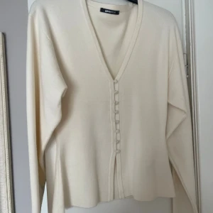 Beige kofta med knappar från Gina Tricot - Snygg beige kofta från Gina Tricot med v-ringning och dekorativa tygklädda knappar framtill. Långärmad och i mjukt stickat material, perfekt för lager-på-lager. Enkel och stilren design som passar till allt. Helt oanvänd.