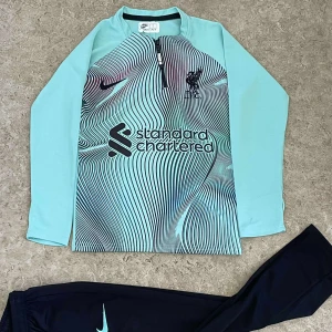 Liverpool långärmad träningströja Nike - Snygg långärmad fotbollströja från Nike för Liverpool FC i ljusblått med svart grafiskt mönster på bröstet. Tröjan har halv dragkedja, klubbmärke och sponsortryck framtill. Tillverkad i lätt och ventilerande Dri-FIT-material.