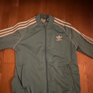 Adidas ljusblå träningsset med dragkedja - Säljer ett snyggt ljusblått Adidas träningsset med klassiska vita ränder längs ärmar och ben. Jackan har dragkedja och Adidas-logga på bröstet, byxorna har resår i midjan och logga på låret. Perfekt för dig som gillar sportig stil.