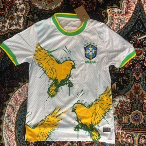 Brasilien fotbollströja  - Snygg vit fotbollströja från Nike med Brasilien CBF-logga på bröstet. Tröjan har gröna och gula detaljer vid krage och ärmslut samt stora gula fåglar tryckta på framsidan. Perfekt för dig som vill sticka ut på match eller träning.
