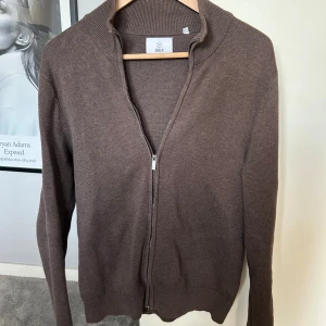 Brun full zip tröja från Bläck - Snygg brun kofta från Bläck med hel dragkedja framtill och ribbade muddar. Tröjan är stickad i ett mjukt material och har en klassisk, enkel design som passar perfekt till jeans eller chinos. Långärmad och med hög krage för extra stilpoäng. Längden på killen i bilden är 183 cm