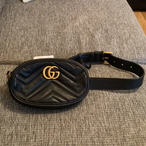 Svart Gucci Marmont i skinn - Snygg svart midjeväska från Gucci i mjukt skinn med guldiga GG-detaljer framtill. Väskan har quiltad yta med chevronmönster och justerbart bälte med guldfärgat spänne. Perfekt storlek för det viktigaste och stängs med dragkedja. Fungerar lika bra att hänga tvärs över axeln eller rakt från axeln. Köpt på Vestiaire Collective men aldrig använd. Som ny och har även genomgått äktighetsintyg. Bredd 18 cm, Höjd 11 cm, Djup 5 cm.