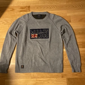 Grå stickad tröja Morris 1968 M - Grå stickad tröja från Morris med broderad patch framtill där det står 'Morris 1968' och en brittisk flagga. Tröjan har rund hals, långa ärmar och är tillverkad i 100% bomull. Perfekt för dig som gillar klassisk och stilren design. 