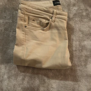 Beiga chinos från J.Lindeberg i storlek 34 - Snygga beiga chinos från J.Lindeberg i klassisk rak modell. Byxorna har fem fickor och diskreta detaljer, perfekta för en stilren look. Tillverkade i bomull för en skön känsla och bra passform.