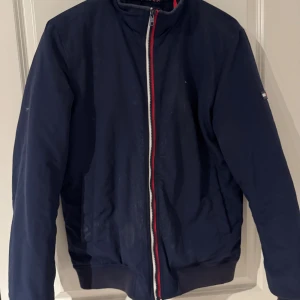 Marinblå bomberjacka Tommy Hilfiger - Snygg marinblå bomberjacka från Tommy Hilfiger Denim med klassisk röd, vit och blå rand vid dragkedjan och muddar. Jackan har ståkrage, två fickor och dragkedja framtill. Perfekt för dig som gillar stilren och sportig look.