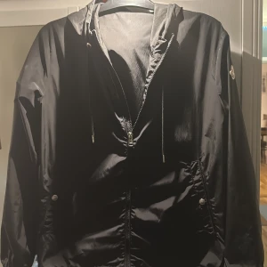 Svart vindjacka från Moncler - Svart vindjacka från Moncler med dragkedja, huva och snörning. Jackan har Moncler-logga på ärmen och snygga röd-vita detaljer på huvan. Tillverkad i glansig polyester och har fickor med tryckknappar. Storlek 2 och självklart äkta! 