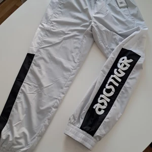 Asics Tiger grå track pants herr L - Snygga sportiga track pants från Asics Tiger i glacier grey med svarta paneler och stor vit logga längs benen. Byxorna har resår i midja och benslut, samt är tillverkade i 100% polyester för en lätt och luftig känsla.