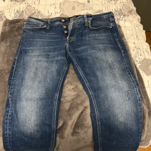 Blå Jack & Jones jeans i modellen Tapered/Mike i storleken 33/30 - Säljer ett par blå jeans från Jack & Jones i storlek 33/30. Jeansen har en tapered passform, klassisk femficksdesign, knäppning med flera knappar och snygga slitningar framtill. Materialet är robust denim med kontrastsömmar och bakfickor med broderad detalj.Den har gått sönder lite vid vänstra knät men vi har sytt den