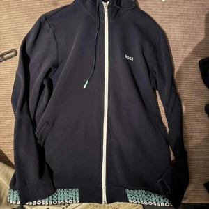 Svart hoodie från BOSS med dragkedja - Snygg svart hoodie från BOSS med vit dragkedja och logga på bröstet. Nederkanten har upprepade BOSS-tryck i vitt och turkost. Tröjan har huva med snörning och långa ärmar, perfekt för en chill och stilren look.