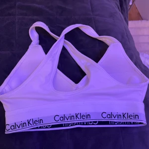 Vit bh  från Calvin Klein - Säljer en vit bh  från Calvin Klein med klassisk logga på det elastiska bandet. Toppen har korsade axelband och ärmlös design, perfekt för en sportig och chill look. Mjuk bomullskänsla och stilren form som sitter skönt mot kroppen!. 🤍