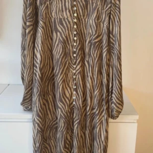 ADOORE Zebramönstrad långklänning  - Snygg långklänning med zebramönster i beige och brunt. Klänningen har långa ärmar, knappar hela vägen framtill och en slits som ger extra edge. Perfekt för dig som vill sticka ut med ett djurmönster och gillar trendiga detaljer.