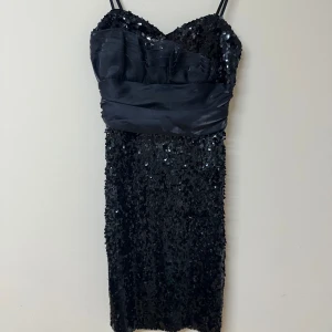 Svart paljettklänning från Byblo - Supersnygg svart kortklänning från Byblo med tunna axelband och glittriga paljetter över hela klänningen. Ovanför midjan finns ett brett band i satinliknande material som ger en cool kontrast. Perfekt för dig som vill sticka ut med en glamorös look.