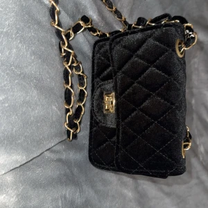 Svart quiltad axelväska i sammet - Snygg svart axelväska i mjuk sammet med quiltat mönster och guldfärgad kedja. Liten och smidig modell med vridlås framtill. Perfekt för dig som vill ha en stilren och trendig accessoar med lyxig känsla.