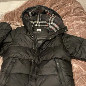 Svart pufferjacka från Burberry - Svart pufferjacka från Burberry med stor huva och klassiskt rutigt foder i grått, vitt och rött. Jackan har dragkedja, knappar och två fickor med dragkedja framtill. Tydlig Burberry-logga på ärmen. Perfekt för kalla dagar och riktigt snygg streetstil.