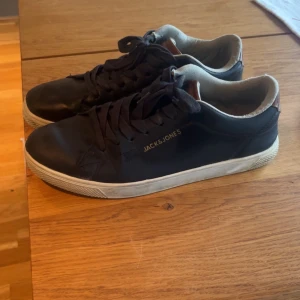 Svarta sneakers från Jack & Jones - Svarta sneakers från Jack & Jones med klassisk låg profil och rund tå. Skorna har snörning och vit platt sula, samt diskret logga på sidan. Ovandelen är i skinn med detaljer i brunt vid hälen och insidan är ljusbeige. Perfekta för en clean och stilren look.