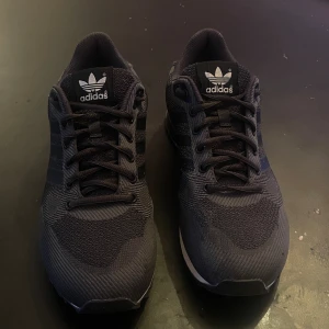 Adidas ZX 750 svarta sneakers - Svarta Adidas ZX 750 sneakers med klassiska tre ränder på sidan och vit sula. Skorna har snörning, rund tå och är tillverkade i syntetmaterial med meshstruktur för extra komfort. Snygg retrostil med Adidas logga på plös och häl. Perfekt för dig som gillar streetwear.