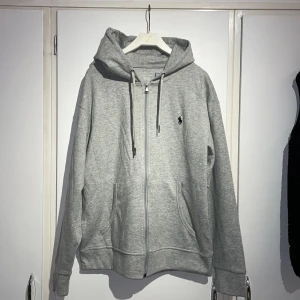 Grå hoodie från Polo Ralph Lauren - Snygg grå hoodie från Polo Ralph Lauren med dragkedja, huva med snörning och klassisk broderad logga på bröstet. Mjuk bomullsblandning och två fickor framtill. Perfekt för chill dagar eller när du vill ha en clean streetstyle. Priset är ej hugget i sten!✌🏽🙌🏻