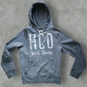 HCO wave devils hoodie - Säljer en grå hoodie från Hollister med stort vitt HCO-tryck och texten 'Wave Devils' på bröstet. Tröjan har huva med snörning och en klassisk känguruficka framtill. Perfekt för chill dagar och snygg streetstil.