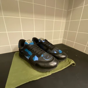 Valentino Rockrunner sneakers svart/blå - Säljer ett par Valentino Rockrunner sneakers i svart och blått med coolt kamouflagemönster. Skorna har snörning, platt sula och detaljer i både skinn och mocka. Perfekt för dig som vill sticka ut med ett lyxigt streetwear vibe.