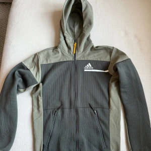 Grön hoodie från Adidas - Tröjan har sportig look med vit Adidas-logga och rand på bröstet. Tillverkad i mjuk polyester med strukturerad yta och detaljer i olika gröna nyanser. Mycket bra skick, storlek S men passar även en lite mindre M.