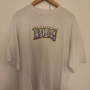 Vit Notorious BIG t-shirt - Vit t-shirt med stort tryck av The Notorious BIG på både fram- och baksidan. Framsidan har texten 'BIG' i blått och svart, baksidan har ett stort motiv med ansikte, text och stadssilhuett. Skön passform och klassisk rund hals. Står storlek S men är oversize så rekommenderar dem som har på sig storlek M