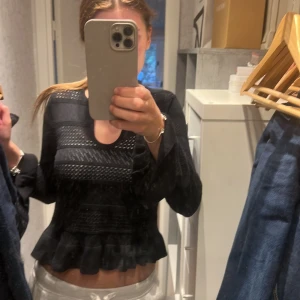 Svart blus med volang och hålmönster - Supersnygg svart blus med långärmade ärmar och genomgående hålmönster. Toppen har en rundad halsringning och volangkant nedtill som ger en feminin touch. Materialet är stickat och känns mjukt mot huden. Perfekt för dig som gillar detaljer och vill ha något extra. till vardag men också fest, använd fåtal gånger