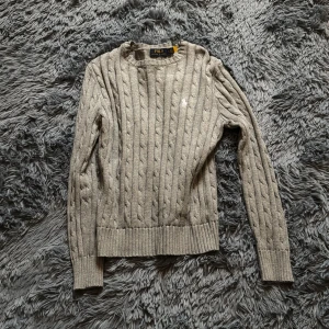 Grå stickad tröja Polo Ralph Lauren - Mycket minde i storleken, det står M men passar bäst xs. Kabelstickad grå tröja från Polo Ralph Lauren med klassisk rund halsringning och broderad logga på bröstet. Tröjan har långa ärmar och breda ribbade muddar vid ärmslut och nederkant. Perfekt för lager-på-lager under kyliga dagar. 