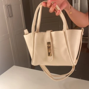 Beige axelväska med gulddetalj - Snygg beige axelväska i syntetmaterial med stilren design och guldfärgat spänne framtill. Väskan har både handtag och axelrem, vilket gör den enkel att bära på olika sätt. Perfekt för dig som vill ha en clean och modern look. Aldrig använd 