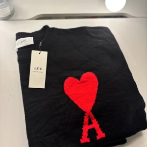 Svart stickad tröja med rött hjärta från Ami - Svart stickad tröja från Ami Alexandre Mattiussi med ett stort rött hjärta och bokstaven A på bröstet. Tröjan är gjord i ekologisk bomull och har en klassisk rund halsringning. Perfekt för dig som gillar stilrena plagg med en twist.